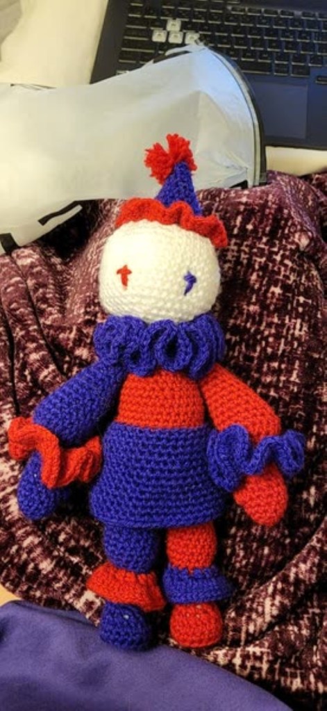 A purple and red crochet clown with ruffles and a white face